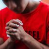 Lähikuva nuoresta miehestä rukoilemassa. Hänen kasvonsa ovat sumeat. Yllään hänellä on punainen t-paita.