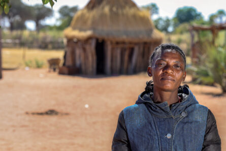 Afrikkalainen nainen seisoo kuvan etualalla. Taustalla perinteinen namibialainen pihapiiri, pieni asuinrakennus ja aitaus.