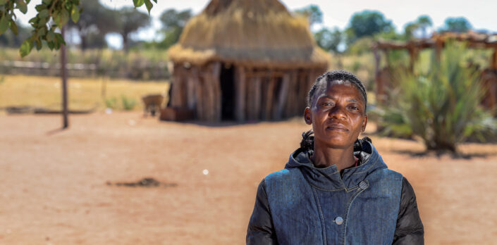 Afrikkalainen nainen seisoo kuvan etualalla. Taustalla perinteinen namibialainen pihapiiri, pieni asuinrakennus ja aitaus.