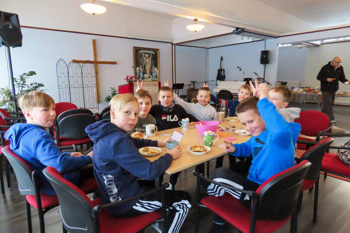 Kuvassa on kymmenkunta alakouluikäistä poikaa pöydän ympärillä välipalaa nauttimassa. Pojat ovat kääntyneet kameraan päin. Moni heistä hymyilee. Useampi on tehnyt kaverin pään taakse niin sanotut pupunkorvat.