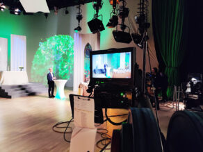 Kuvassa on etualalla televisiokamera, joka kuvaa taustalla puhetta pitävää Kari Hämäläistä, Jyväskylän vapaaseurakunnan pastoria. Studiossa on hämärää. Lattia on vaalea ja lavasteet valkoiset. Seinälle on heijastettu vihreä puu ja vihreää valoa.