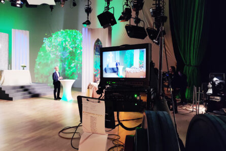 Kuvassa on etualalla televisiokamera, joka kuvaa taustalla puhetta pitävää Kari Hämäläistä, Jyväskylän vapaaseurakunnan pastoria. Studiossa on hämärää. Lattia on vaalea ja lavasteet valkoiset. Seinälle on heijastettu vihreä puu ja vihreää valoa.