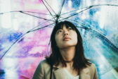 Aasialainen nuori nainen seisoo sateenvarjon alla katsoen totisena yläviistoon. Kuvan tausta on sini- ja purppurasävyinen.
