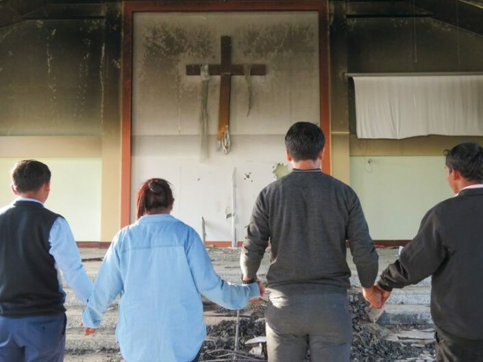 Neljä ihmistä selin, käsi kädessä, kasvot kohti seinällä olevaa ristiä. Huoneessa jälkiä tuhopoltosta.