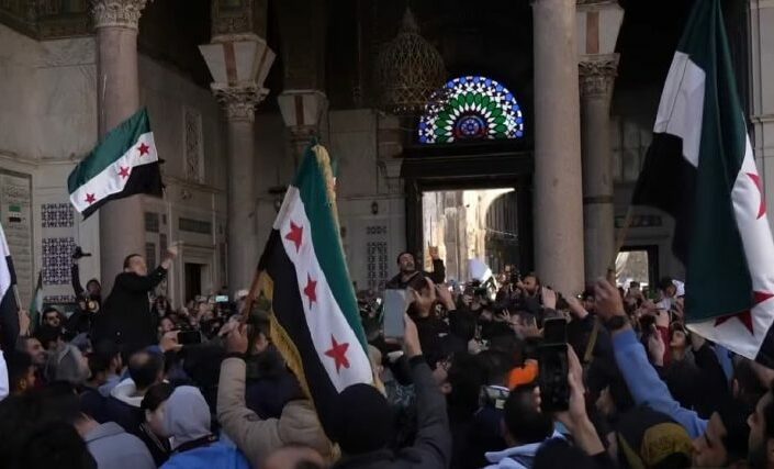 Valokuva jossa joukko iloitsevia ihmisiä ja Syyrian lippuja moskeijassa.