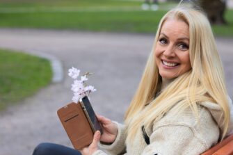 Nainen katsoo kameraan puhelin kädessään ja hymyilee