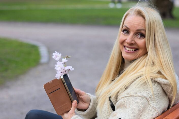 Nainen katsoo kameraan puhelin kädessään ja hymyilee