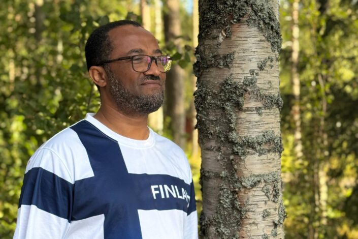 Mies seisoo Koivu-puun vieressä ja hymyilee.