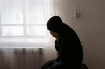 Tummaan pukuun ja huiviin pukeutunut nainen polvillaan lattialla ikkunan vieressä, kasvot kätkettyinä käsiinsä.