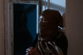 Nainen, jonka kasvoista osa on peitetty huivilla, istuu ikkunalla kädet ristissä ja katsoo ikkunasta ulos.