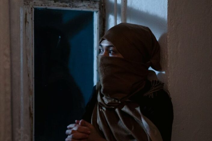 Nainen, jonka kasvoista osa on peitetty huivilla, istuu ikkunalla kädet ristissä ja katsoo ikkunasta ulos.