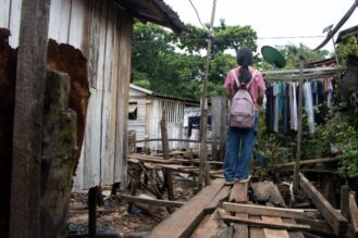 Tyttö reppu selässään, selin kameraan, laudoista kyhätyn sillan päällä, ympärillä paalujen päällä olevia puurakennuksia.