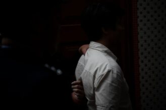 Kädet tarttuvat miehen niskaan ja selkään takaapäin, kummankin ihmisen kasvot jäävät pimeään.