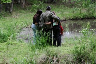Mies ja kolme lasta selin kameraan lammen rannassa, kädet kiedottuina toistensa kauloille.
