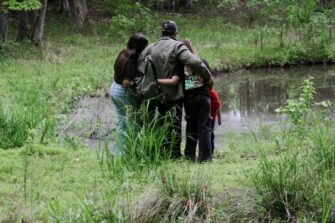 Mies ja kolme lasta selin kameraan lammen rannassa, kädet kiedottuina toistensa kauloille.