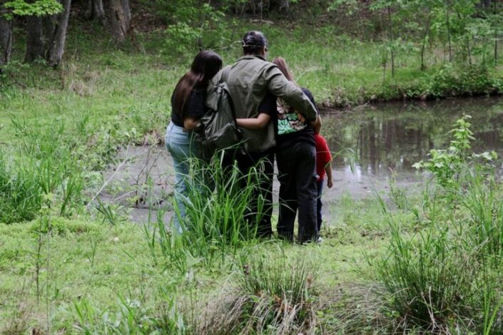 Mies ja kolme lasta selin kameraan lammen rannassa, kädet kiedottuina toistensa kauloille.