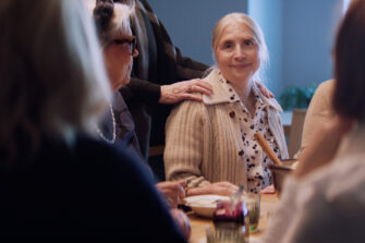 Nainen istuu ruokapöydässä toisten seurassa. Hän hymyilee lempeästi kameraan, ja toinen henkilö laskee rohkaisevasti kätensä hänen olkapäälleen. Tilanne välittää yhteisöllisyyttä ja tukea.