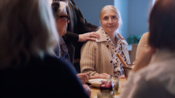 Nainen istuu ruokapöydässä toisten seurassa. Hän hymyilee lempeästi kameraan, ja toinen henkilö laskee rohkaisevasti kätensä hänen olkapäälleen. Tilanne välittää yhteisöllisyyttä ja tukea.