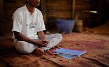 Mies istuu matolla edessään pari vihkoa. Kasvot on rajattu kuvan ulkopuolelle.