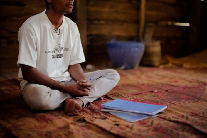 Mies istuu matolla edessään pari vihkoa. Kasvot on rajattu kuvan ulkopuolelle.