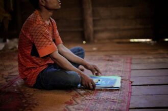 Mies istuu lattialla edessään kasa koulukirjoja ja vihkoja.