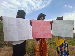 Kolme afrikkalaista nuorta naista käsissään suuret julisteet, joihin on kirjoitettu englanniksi tekstejä, joissa luvataan rukoilla Leah Sharibun puolesta.