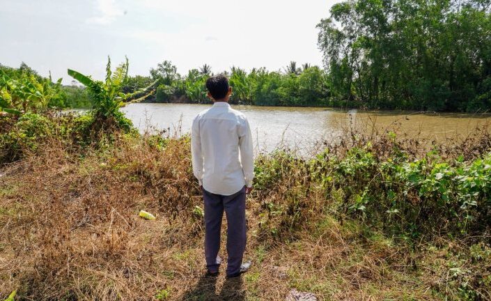 Mies seisoo joen rannalla selin kameraan.