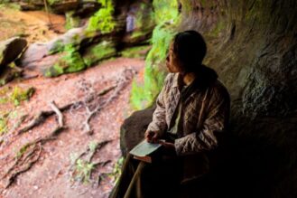 Aasialainen nainen istuu suuren puun juurella Raamattu sylissään.