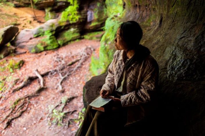 Aasialainen nainen istuu suuren puun juurella Raamattu sylissään.