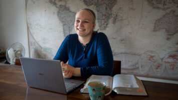 Susanna Sorila, vaaleahiuksinen nainen sinisessä paidassa, istuu pöydän äärellä edessään avoin Raamattu ja tietokone.