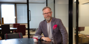 Mies toimistossa katsoo suoraan kameraan ja hymyilee