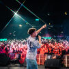Rap-artisti Amos esiintyy mikrofoni kädessään ja toinen käsi kohotettuna. Taustalla näkyy suuri yleisö ja puhelinten valoja.