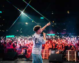 Rap-artisti Amos esiintyy mikrofoni kädessään ja toinen käsi kohotettuna. Taustalla näkyy suuri yleisö ja puhelinten valoja.