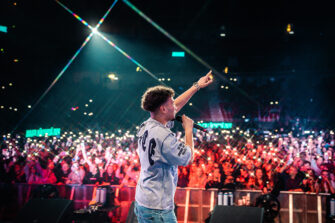 Rap-artisti Amos esiintyy mikrofoni kädessään ja toinen käsi kohotettuna. Taustalla näkyy suuri yleisö ja puhelinten valoja.
