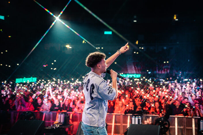 Rap-artisti Amos esiintyy mikrofoni kädessään ja toinen käsi kohotettuna. Taustalla näkyy suuri yleisö ja puhelinten valoja.